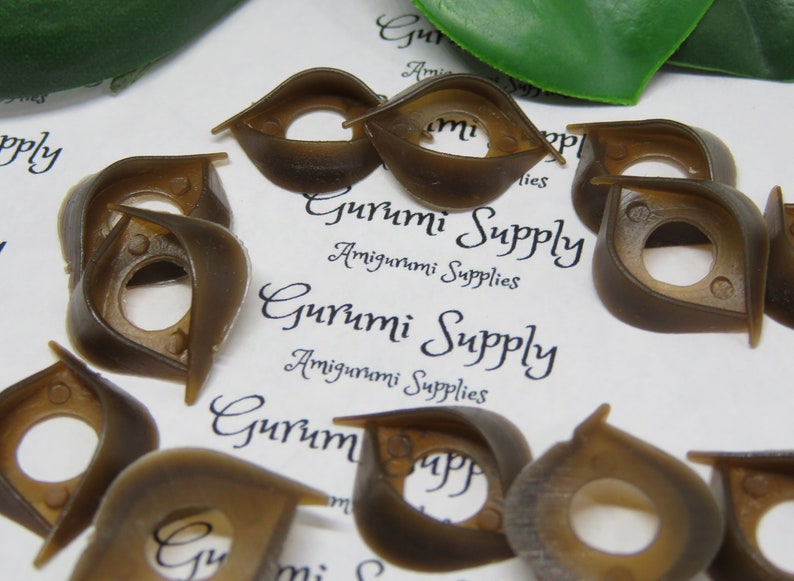 May include: A collection of brown plastic safety eyes for amigurumi projects. The eyes have a unique shape with a central hole and a curved, wing-like design. The text "Gurumi Supply" and "Amigurumi Supplies" is visible.