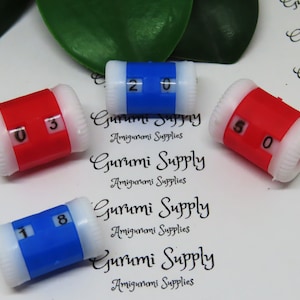 May include: Four plastic stitch counter rings. Two are blue with white numbers and two are red with white numbers. The numbers on the rings are 03, 18, 20, and 50.