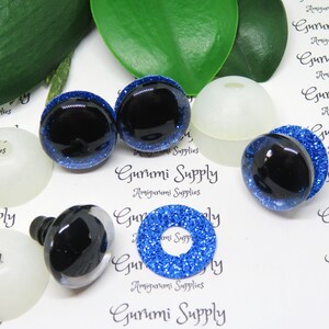 Peut inclure: Quatre yeux de sécurité noirs et bleus avec des paillettes, deux rondelles d'yeux de sécurité en plastique blanc et un cercle de paillettes bleu. Gurumi Supply Amigurumi Supplies est imprimé sur le fond blanc.