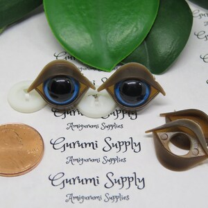 May include: Brown plastic doll eyes with black pupils and blue irises. The eyes are shaped like an almond and have a brown frame. The image also includes a penny and the text "Gurumi Supply Amigurumi Supplies".