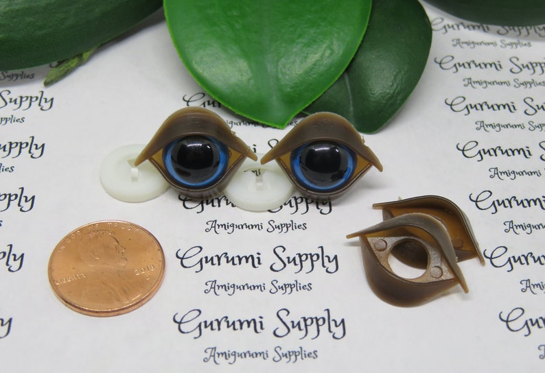 May include: Brown plastic doll eyes with black pupils and blue irises. The eyes are shaped like an almond and have a brown frame. The image also includes a penny and the text "Gurumi Supply Amigurumi Supplies".