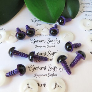May include: A collection of purple and black safety eyes for amigurumi projects. The eyes have a glossy finish and screw-on backs, with white plastic washers. The text "Gurumi Supply" is printed on the background.