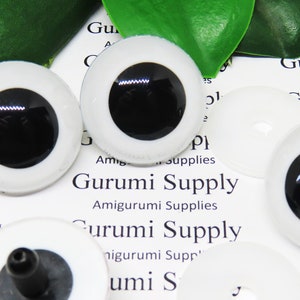 Puede incluir: Tres ojos de seguridad de plástico blanco con pupilas negras. Los ojos están sobre un fondo blanco con el texto "Gurumi Supply" impreso en él.