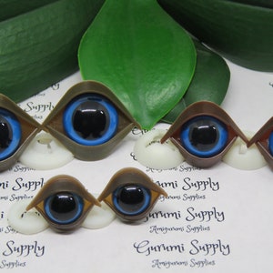 May include: A collection of doll eyes in various shades of brown and tan, with vibrant blue irises and black pupils. The eyes are set on white bases, with a green leaf in the background. The text "Gurumi Supply" is visible.