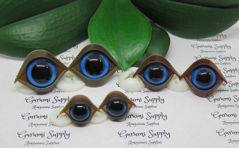 May include: A collection of doll eyes in various shades of brown and tan, with vibrant blue irises and black pupils. The eyes are set on white bases, with a green leaf in the background. The text "Gurumi Supply" is visible.