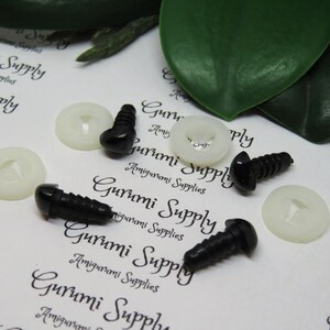 May include: A close-up of six black plastic safety eyes with white plastic washers. The eyes are on a white surface with the text "Gurumi Supply" printed on it.