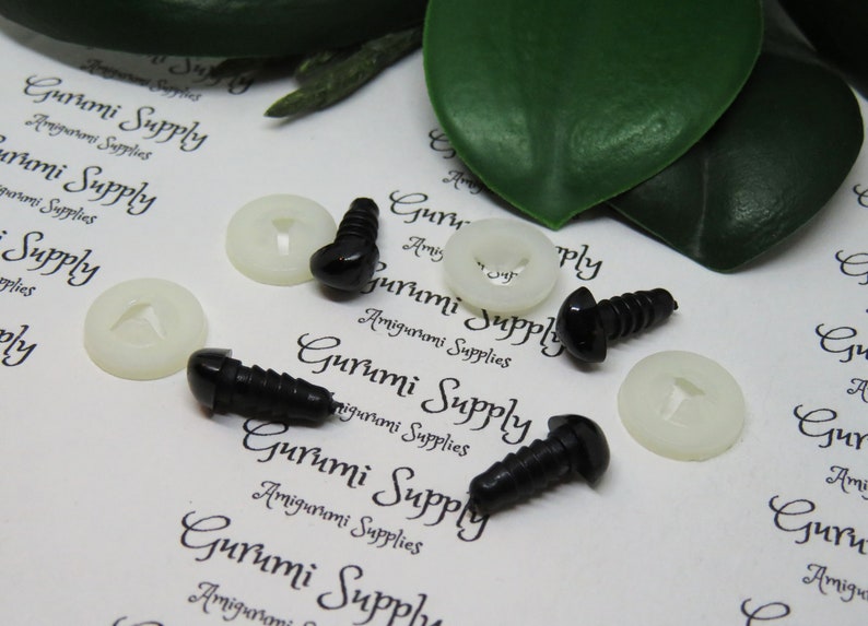May include: A close-up of six black plastic safety eyes with white plastic washers. The eyes are on a white surface with the text "Gurumi Supply" printed on it.