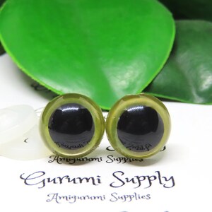 Puede incluir: Dos ojos de seguridad verdes con pupilas negras, utilizados para proyectos de amigurumi. Los ojos est&aacute;n sobre un fondo blanco con una arandela blanca detr&aacute;s de cada ojo.