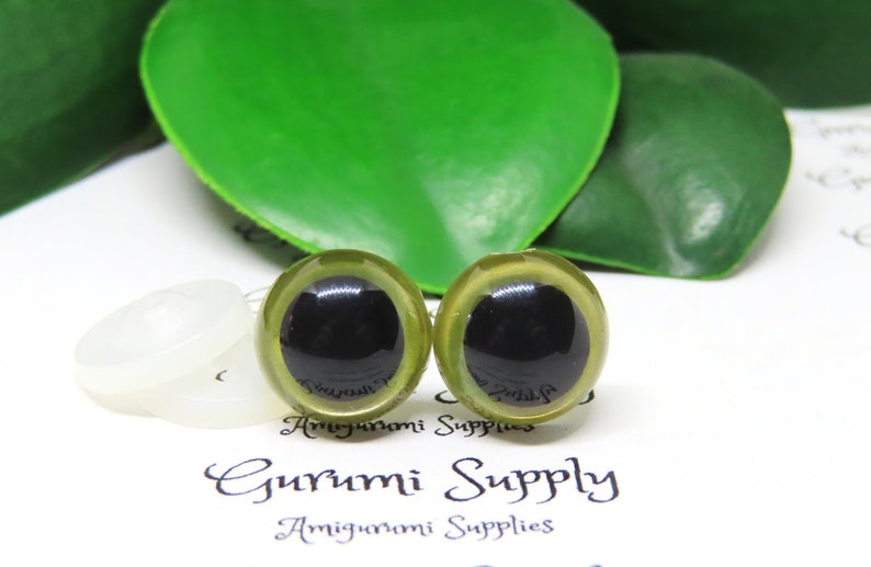 Puede incluir: Dos ojos de seguridad verdes con pupilas negras, utilizados para proyectos de amigurumi. Los ojos est&aacute;n sobre un fondo blanco con una arandela blanca detr&aacute;s de cada ojo.