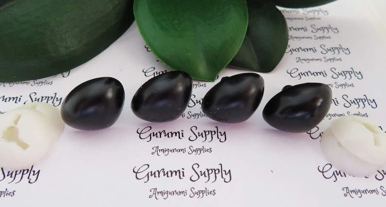 May include: Four black plastic safety eyes for amigurumi projects. The eyes are oval shaped and have a glossy finish. The eyes are on a white background with the text "Gurumi Supply Amigurumi Supplies" printed on it.