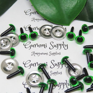 May include: A close-up of a collection of green plastic safety eyes with black plastic backs. The eyes are scattered on a white surface with the text "Gurumi Supply" printed on it. The eyes are used for amigurumi projects.