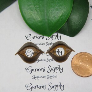 May include: Two brown plastic amigurumi safety eyes with a copper penny for size comparison. The eyes have a rounded shape with a flat top and a circular opening in the center. The text "Gurumi Supply" is printed on the background.