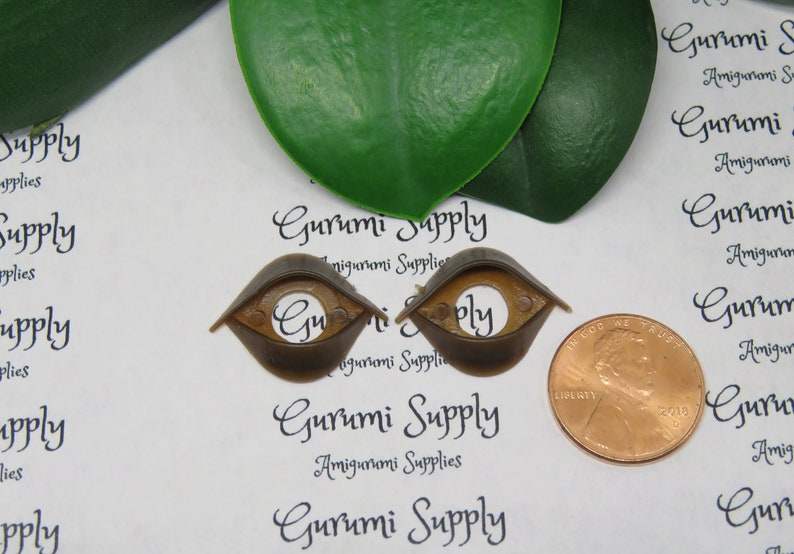 May include: Two brown plastic amigurumi safety eyes with a copper penny for size comparison. The eyes have a rounded shape with a flat top and a circular opening in the center. The text "Gurumi Supply" is printed on the background.