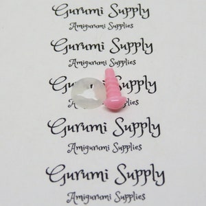 Pu&ograve; includere: Primo piano di occhi di sicurezza in plastica rosa e bianca per progetti di amigurumi. Gli occhi sono su uno sfondo bianco con il testo "Gurumi Supply Amigurumi Supplies" ripetuto pi&ugrave; volte.