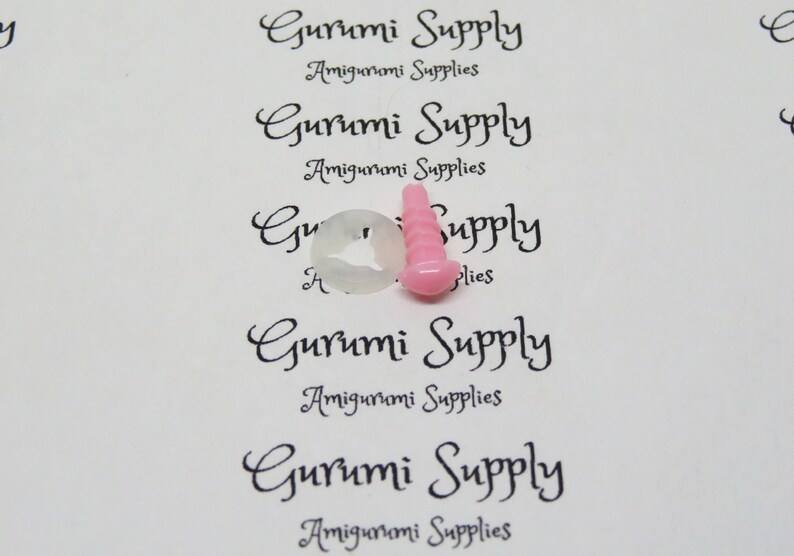 Pu&ograve; includere: Primo piano di occhi di sicurezza in plastica rosa e bianca per progetti di amigurumi. Gli occhi sono su uno sfondo bianco con il testo "Gurumi Supply Amigurumi Supplies" ripetuto pi&ugrave; volte.