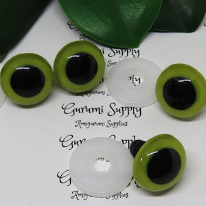 May include: Close-up of six green plastic safety eyes with black pupils. The eyes are arranged in a semi-circle on a white surface. The text "Gurumi Supply Amigurumi Supplies" is visible in the background.