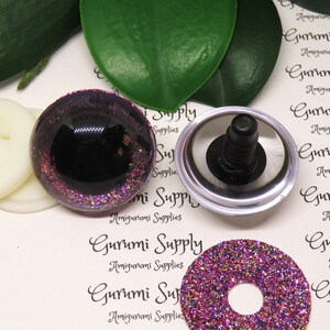 May include: A set of purple glitter safety eyes for amigurumi projects. The eyes are 12mm in diameter and have a black pupil. The set includes two eyes, two washers, and two plastic backings.