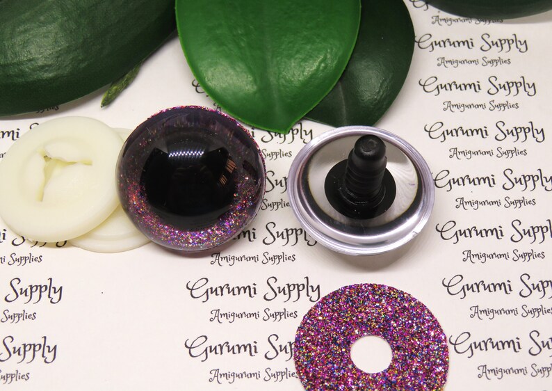 May include: A set of purple glitter safety eyes for amigurumi projects. The eyes are 12mm in diameter and have a black pupil. The set includes two eyes, two washers, and two plastic backings.