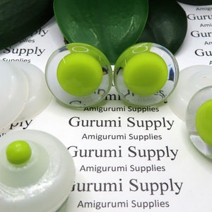 Op de afbeelding: Close-up van vier sets groene en transparante plastic veiligheidsogen voor amigurumi-projecten. De ogen zijn gerangschikt op een wit oppervlak met de tekst "Gurumi Supply" erop gedrukt.