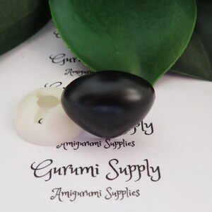 May include: A black plastic safety eye for amigurumi projects. The eye is round and has a glossy finish. The eye is sitting on a white background with the text "Gurumi Supply" printed on it.