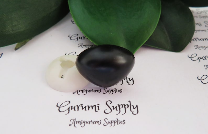 May include: A black plastic safety eye for amigurumi projects. The eye is round and has a glossy finish. The eye is sitting on a white background with the text "Gurumi Supply" printed on it.