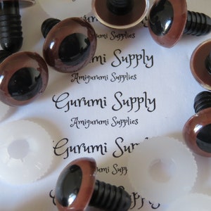 Op de afbeelding: Bruine plastic veiligheidsogen met zwarte pupillen en een zwarte plastic ring. De ogen zijn bevestigd aan een zwarte plastic pen. De ogen liggen op een wit oppervlak met de tekst "Gurumi Supply Amigurumi Supplies" erop gedrukt.