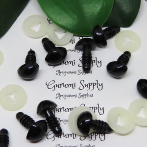 Puede incluir: Ojos de seguridad de plástico negro con tornillo en la parte posterior, para usar en amigurumi y otras manualidades. Los ojos están esparcidos sobre una superficie blanca con arandelas de plástico blanco.