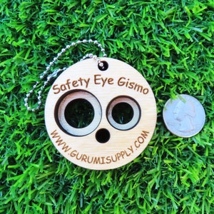 May include: A wooden safety eye tool with two different sized holes for inserting safety eyes. The tool is attached to a silver chain and has the text "Safety Eye Gismo" and "www.gurumisupply.com" engraved on it.