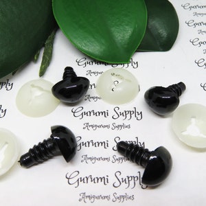May include: Close-up of black plastic safety eyes with screw backs and white plastic washers. The eyes are arranged on a white surface with the text "Gurumi Supply Amigurumi Supplies" printed on it.
