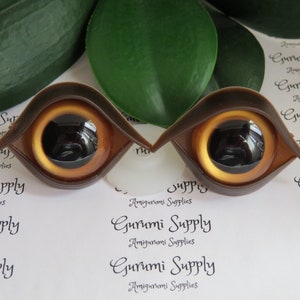 Puede incluir: Dos ojos de mu&ntilde;eca marrones con un iris amarillo dorado y una pupila negra. Los ojos son de pl&aacute;stico y est&aacute;n dise&ntilde;ados para amigurumi u otros proyectos de manualidades. El texto "Gurumi Supply" es visible.