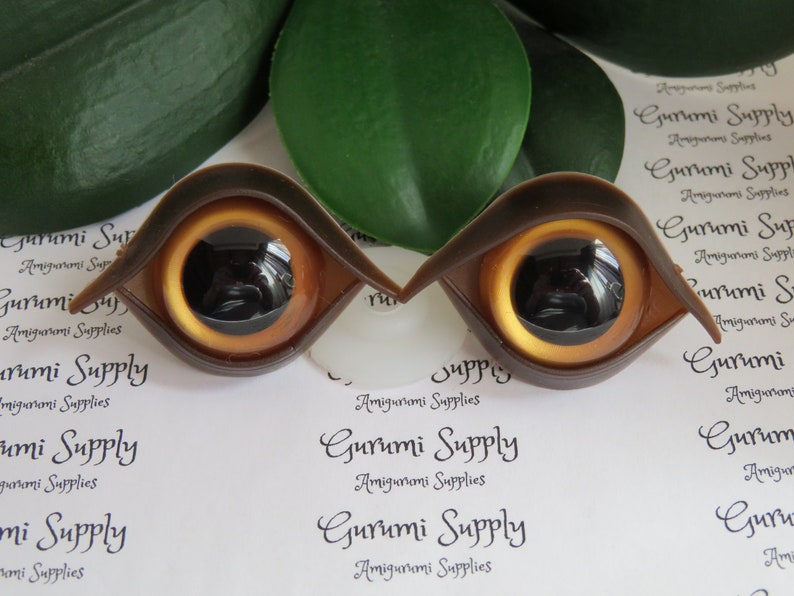Puede incluir: Dos ojos de mu&ntilde;eca marrones con un iris amarillo dorado y una pupila negra. Los ojos son de pl&aacute;stico y est&aacute;n dise&ntilde;ados para amigurumi u otros proyectos de manualidades. El texto "Gurumi Supply" es visible.
