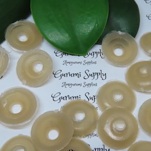Puede incluir: Un conjunto de ojos de seguridad beige en forma de rosquilla para amigurumi. Los ojos están hechos de un material translúcido y tienen un agujero en el centro. El texto "Gurumi Supply Amigurumi Supplies" es visible en el fondo.