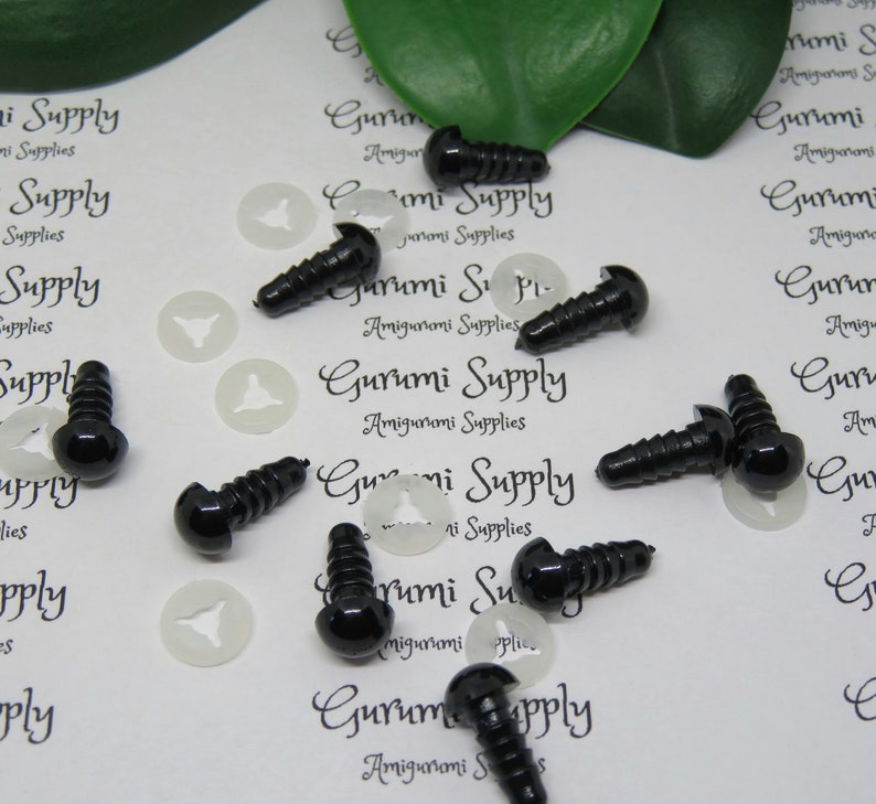 May include: A close-up of black plastic safety eyes with a screw back, used for amigurumi projects. The eyes are scattered on a white surface with white plastic washers.