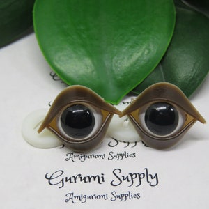 May include: Two brown doll eyes with black pupils and white irises are displayed. The eyes have a unique shape, with a curved, eyelid-like structure. The background includes green leaves and the text "Gurumi Supply Amigurumi Supplies."