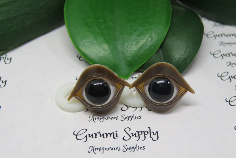 May include: Two brown doll eyes with black pupils and white irises are displayed. The eyes have a unique shape, with a curved, eyelid-like structure. The background includes green leaves and the text "Gurumi Supply Amigurumi Supplies."