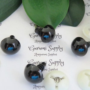 May include: Four black plastic safety eyes with blue pupils, used for amigurumi projects. The eyes are on a white background with the text "Gurumi Supply Amigurumi Supplies" printed on it.