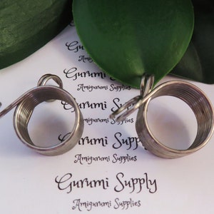 Könnte beinhalten: Zwei silberfarbene Maschenmarkierer aus Metall mit Spiraldesign. Die Markierer haben oben eine Schlaufe und ein gebogenes Ende. Der Hintergrund umfasst ein grünes Blatt und den Text "Gurumi Supply Amigurumi Supplies".