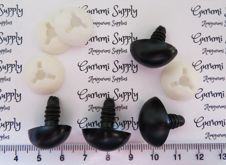 May include: Close-up of black and white plastic safety eyes for amigurumi projects. The eyes are 12mm in diameter and have a screw-on back. The image shows the eyes next to a ruler, with the text "Gurumi Supply" printed on the background.
