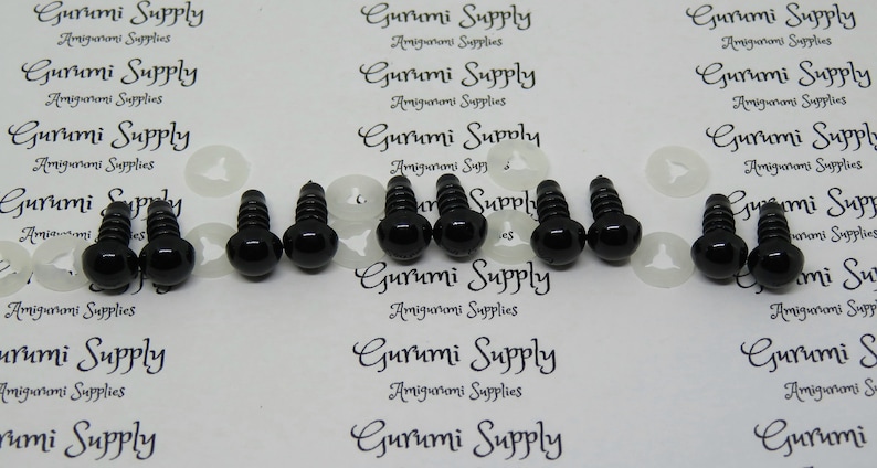 May include: A close-up of eight black plastic safety eyes with screw backs. The eyes are arranged in two rows of four. The eyes are on a white background with the text "Gurumi Supply" printed on it.