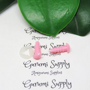 Pu&ograve; includere: Due occhi di sicurezza in plastica rosa per progetti di amigurumi, insieme a una rondella di plastica trasparente. Gli occhi hanno una superficie testurizzata e sono leggermente rialzati. Il testo "Gurumi Supply Amigurumi Supplies" &egrave; stampato sullo sfondo bianco.