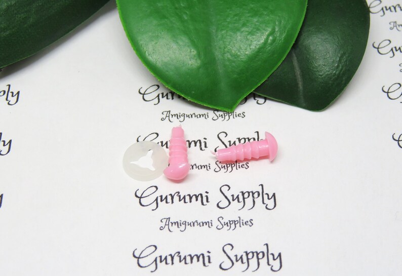 Pu&ograve; includere: Due occhi di sicurezza in plastica rosa per progetti di amigurumi, insieme a una rondella di plastica trasparente. Gli occhi hanno una superficie testurizzata e sono leggermente rialzati. Il testo "Gurumi Supply Amigurumi Supplies" &egrave; stampato sullo sfondo bianco.