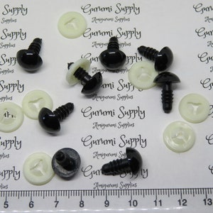 May include: A close-up of black plastic safety eyes with white plastic washers. The eyes are approximately 1 inch in diameter and have a screw-on back. The eyes are arranged on a white surface with a ruler in the bottom of the image.