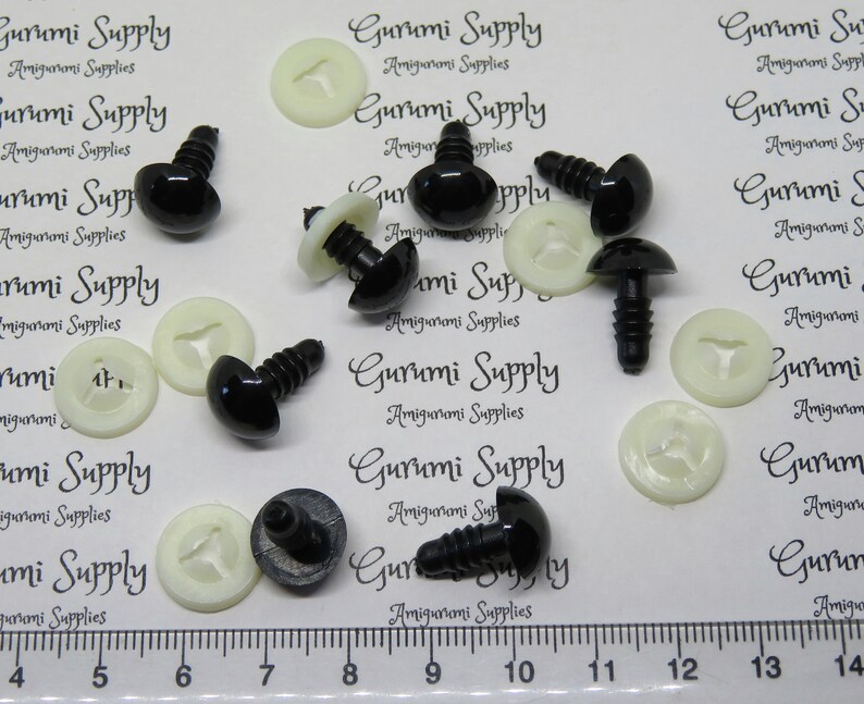 May include: A close-up of black plastic safety eyes with white plastic washers. The eyes are approximately 1 inch in diameter and have a screw-on back. The eyes are arranged on a white surface with a ruler in the bottom of the image.