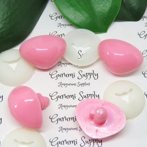 May include: Assortment of pink and white plastic safety eyes for amigurumi projects. The eyes are in various sizes and shapes, including round, oval, and with a flat back.
