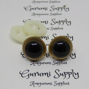 Puede incluir: Dos ojos de seguridad de pl&aacute;stico de color dorado con pupilas negras. Los ojos est&aacute;n sobre un fondo blanco con el texto "Gurumi Supply" impreso en &eacute;l.