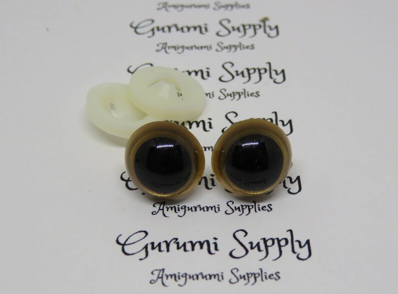 Puede incluir: Dos ojos de seguridad de pl&aacute;stico de color dorado con pupilas negras. Los ojos est&aacute;n sobre un fondo blanco con el texto "Gurumi Supply" impreso en &eacute;l.
