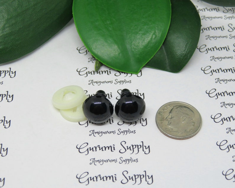 May include: Two black plastic safety eyes for amigurumi projects, next to a white button and a US dime for size comparison. The eyes are 12mm in diameter. The image includes the text "Gurumi Supply Amigurumi Supplies".
