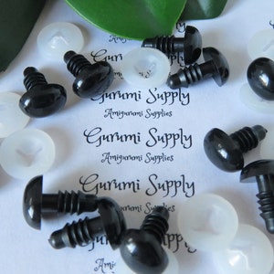 May include: A close-up of black plastic safety eyes with screw backs and white plastic washers. The eyes are arranged on a white surface with the text "Gurumi Supply Amigurumi Supplies" printed on it.