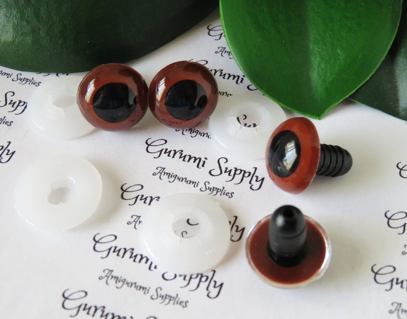Puede incluir: Cuatro ojos de seguridad de pl&aacute;stico marr&oacute;n con pupilas negras y arandelas blancas. Los ojos son para proyectos de amigurumi. El texto "Gurumi Supply Amigurumi Supplies" es visible en el fondo.