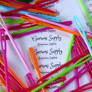 May include: A close-up of a pile of colorful plastic yarn needles. The needles are arranged in a random pattern and are of various lengths. The needles are all the same size and have a small eye at one end. The needles are used for crocheting and knitting.